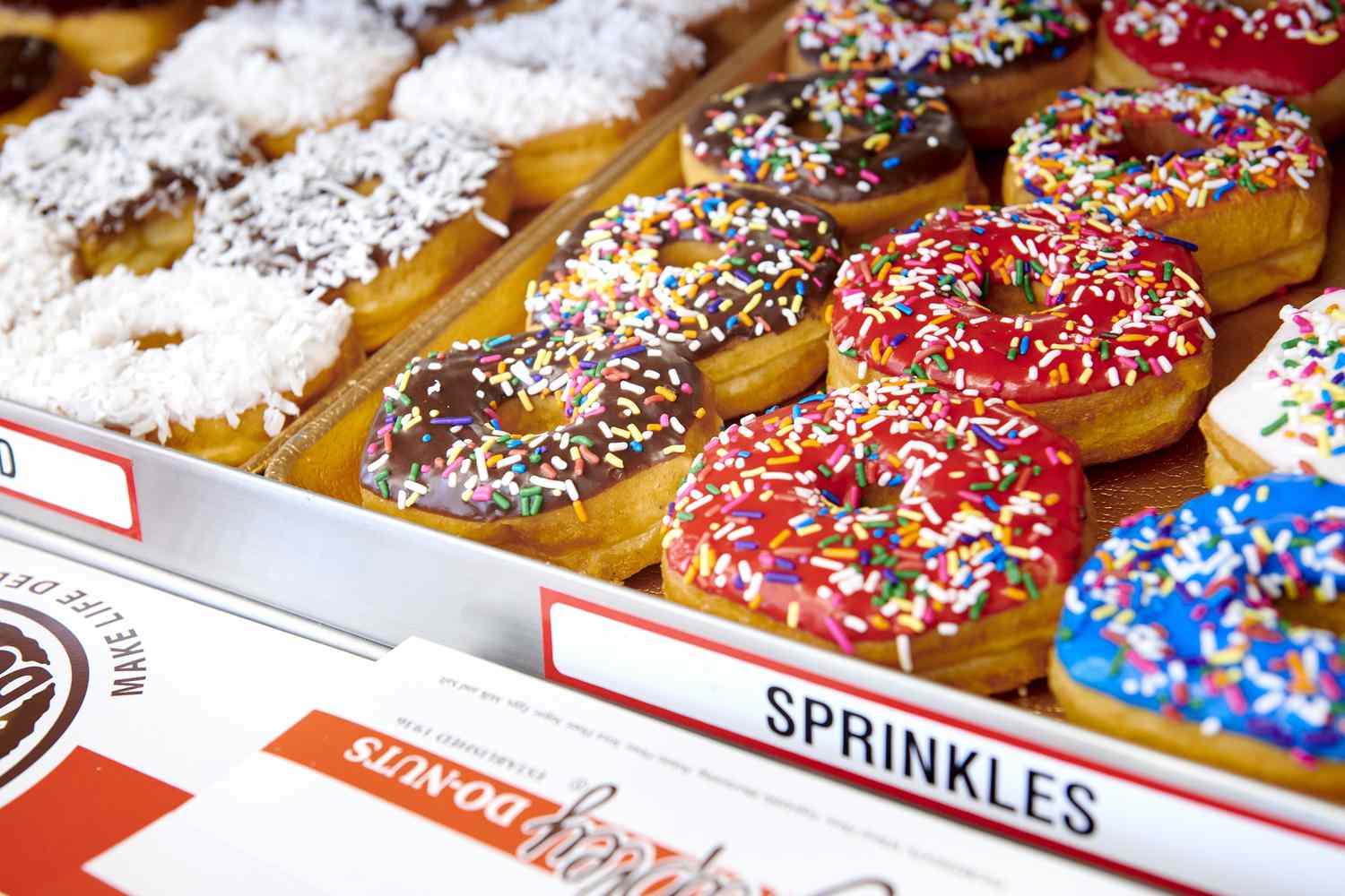 A tray of colorful shipley donuts topped with sprinkles and coconut shavings.